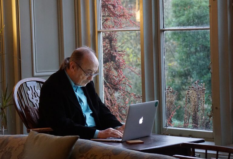 An elderly man sits at a desk, by a window typing on a laptop