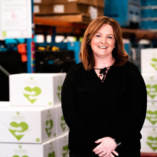 Good360 founder Alison Covington AM standing in a warehouse in front of a stack of Good360 boxes smiling.