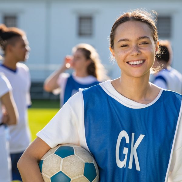 Cheerful soccer player holding a football and looking at camera. Portrait of young woman during training on soccer field. Satisfied high school student holding ball under the arm with her teammmats standing in background.